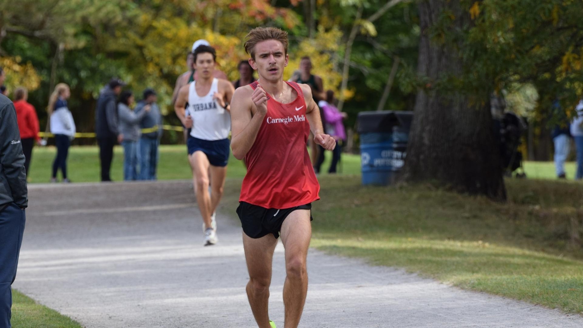 men's cross country runner wearing red jersey and black shorts runs on trail ahead of runners with fans cheering on the left