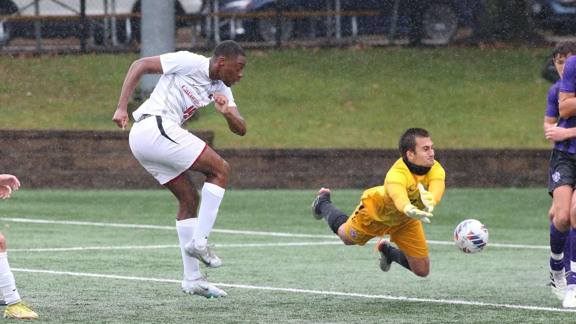 men's soccer player wearing white uniform looks as the keeper wearing yellow dives to stop his shot which hits a defender