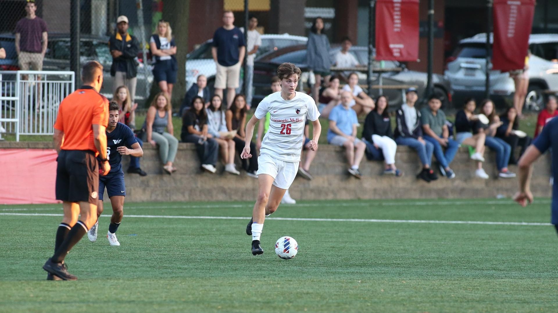 men's soccer player wearing white uniform looks up field to make a pass with a crowd of people in the background watching