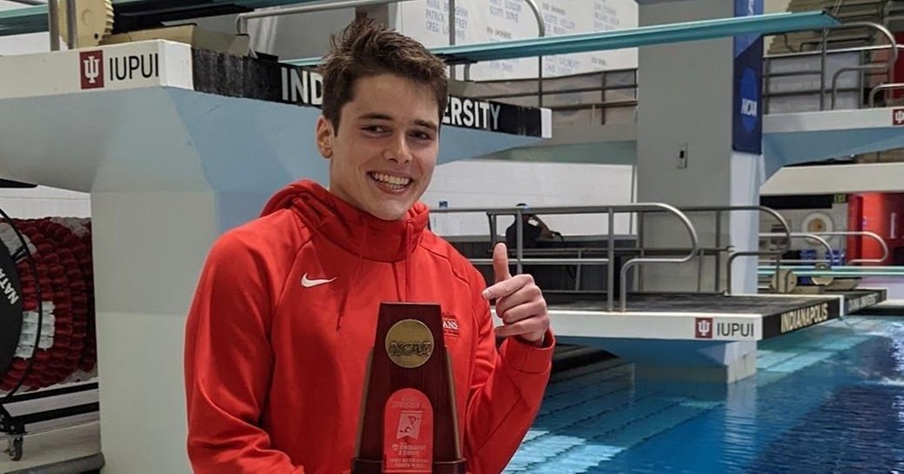 male in red sweatshirt holding NCAA trophy with diving boards in background