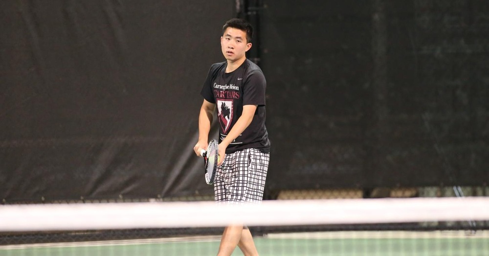 Men's tennis player wearing black top and gray bottoms prepares to serve