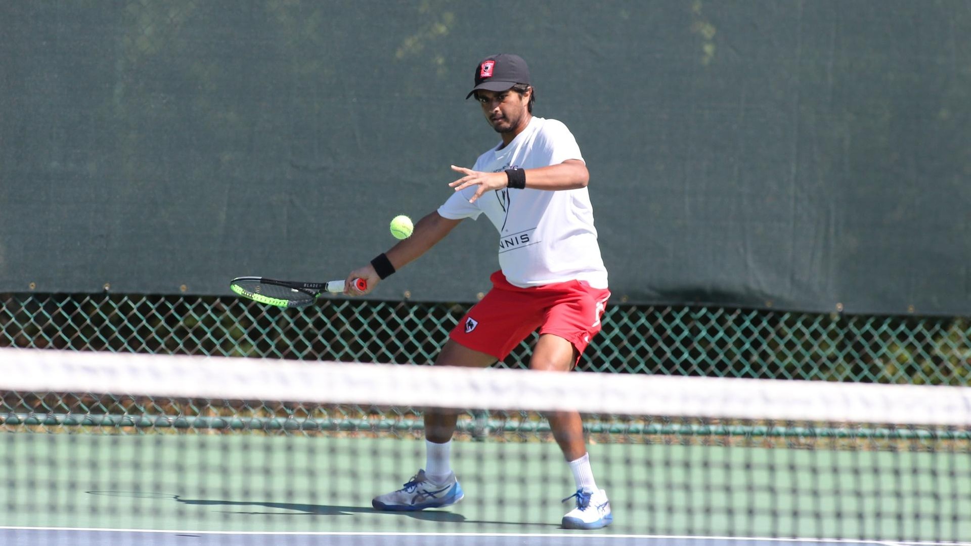 men's tennis player wearing a white shirt, red shorts, and black hat swings at a ball