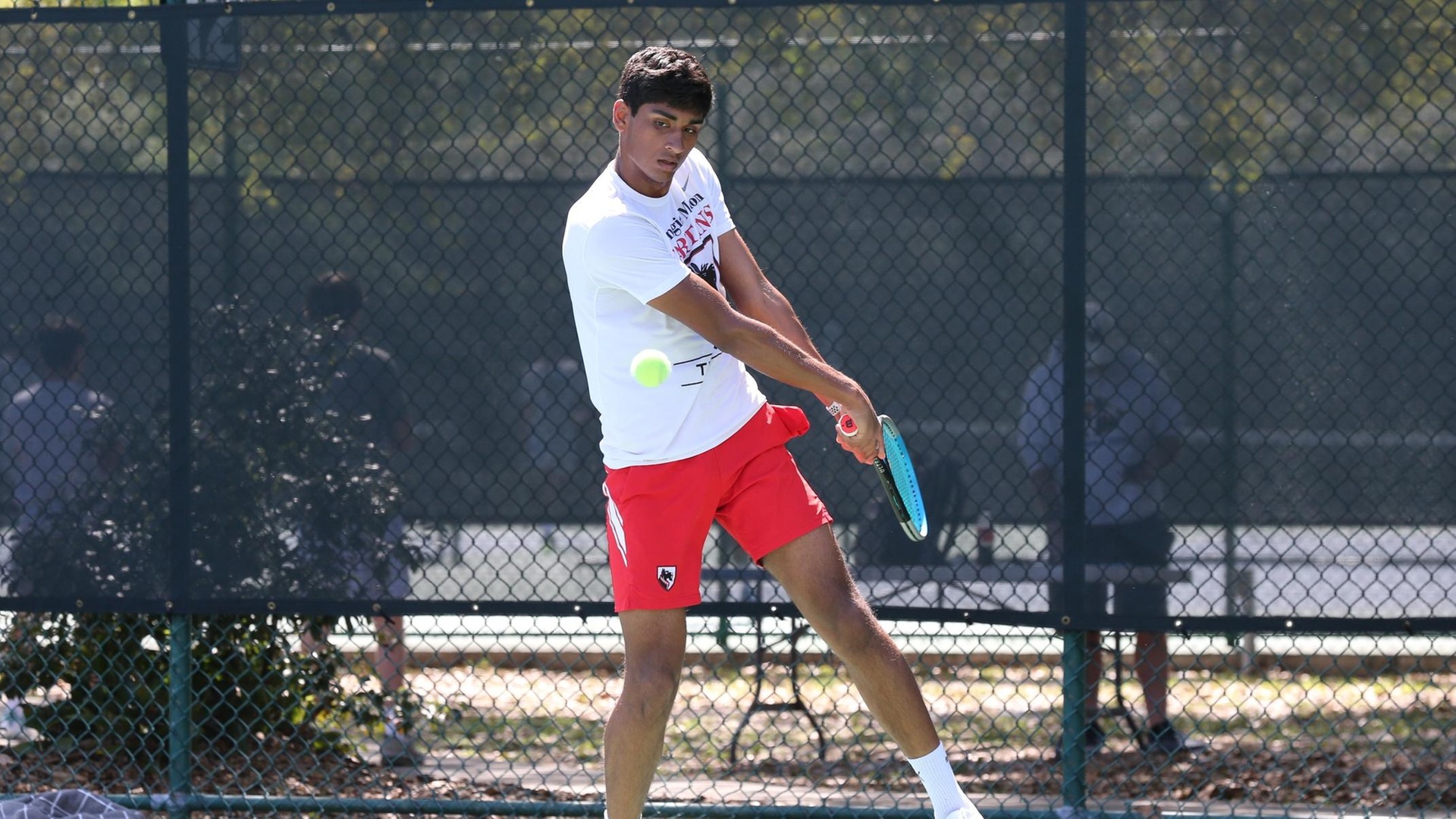 men's tennis player wearing a white shirt and red shorts swinging racquet at the ball