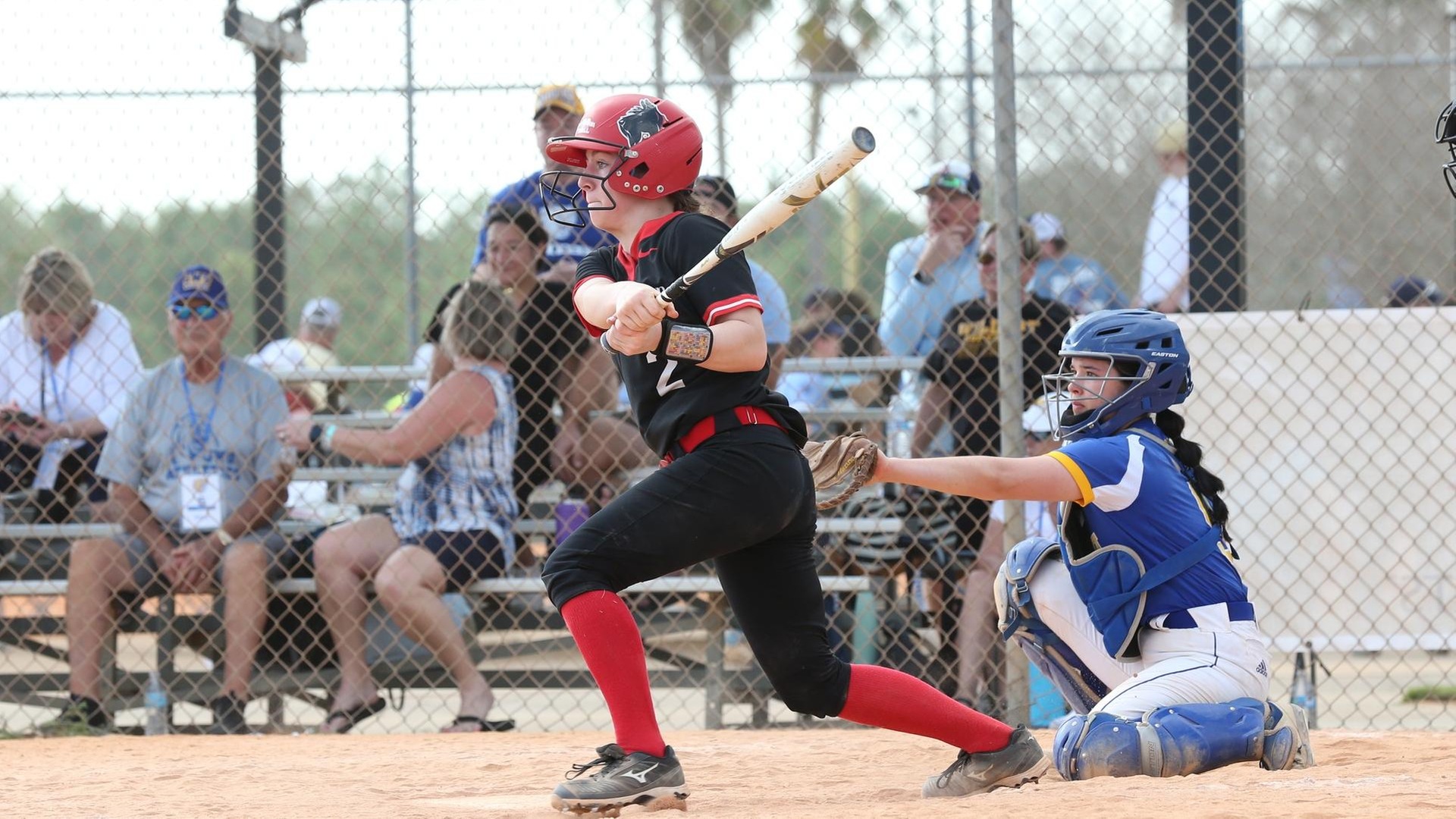 women's softball player wearing a black uniform with a red helmet swings through a pitch