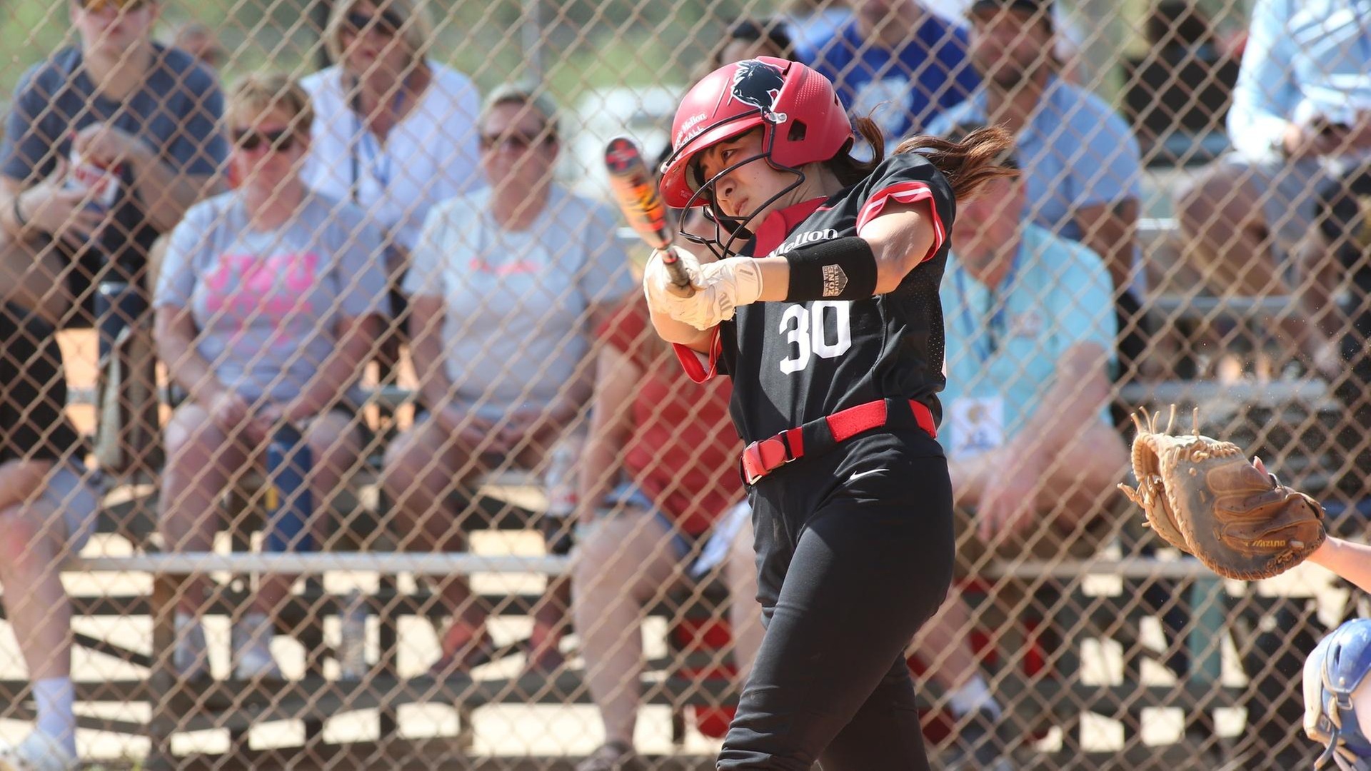 softball player wearing black and red uniform swings at the plate