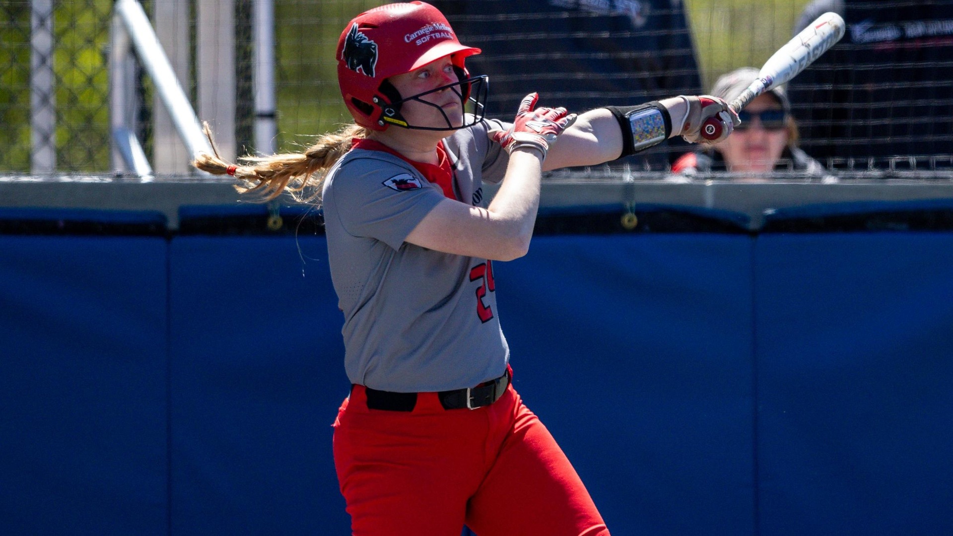 women's softball player wearing a gray jersey and a red helmet takes a swing