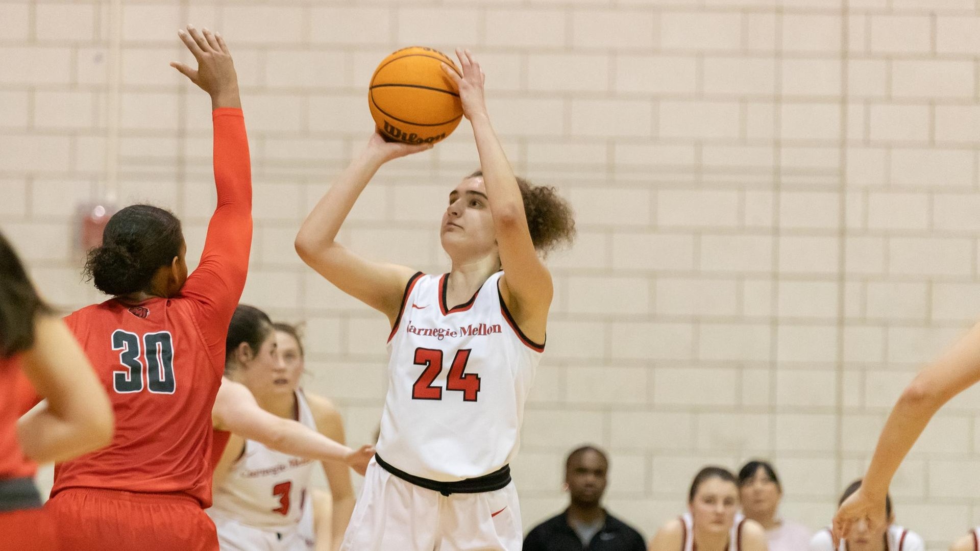 women's basketball player wearing a white uniform takes a shot over the arm of a defender wearing a red uniform