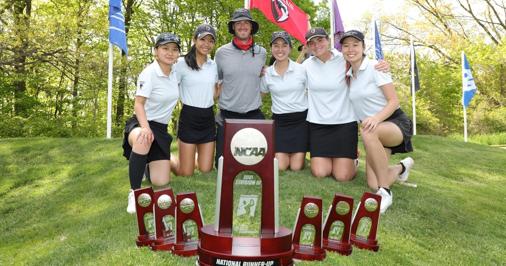 Five women and one man kneeling in grass with trees and a red flag in background with NCAA trophies in the front