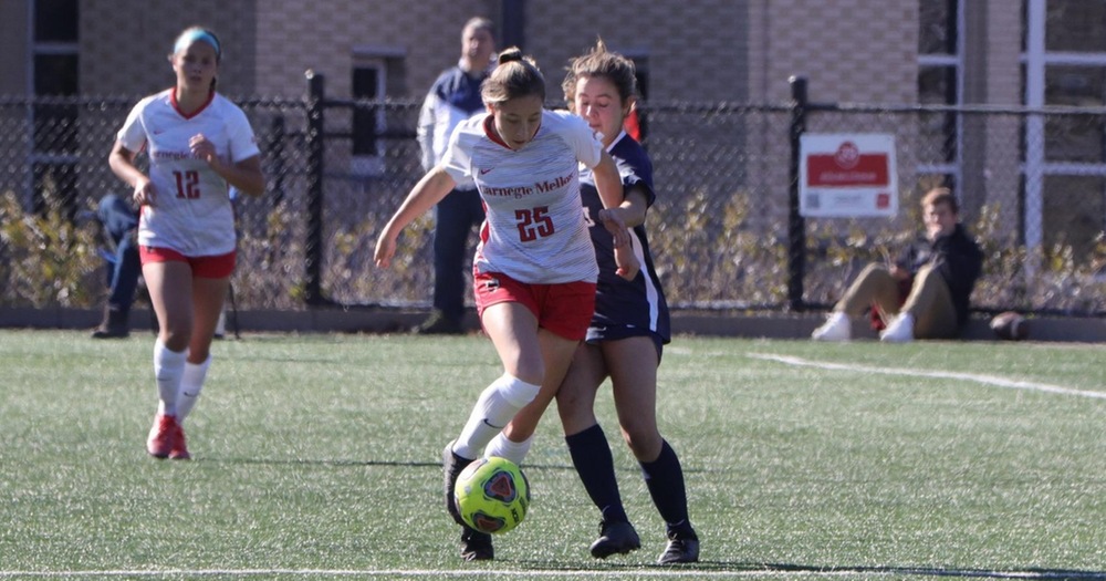 women's soccer player wearing white shirt and red shorts dribbles forward with a defender on her back