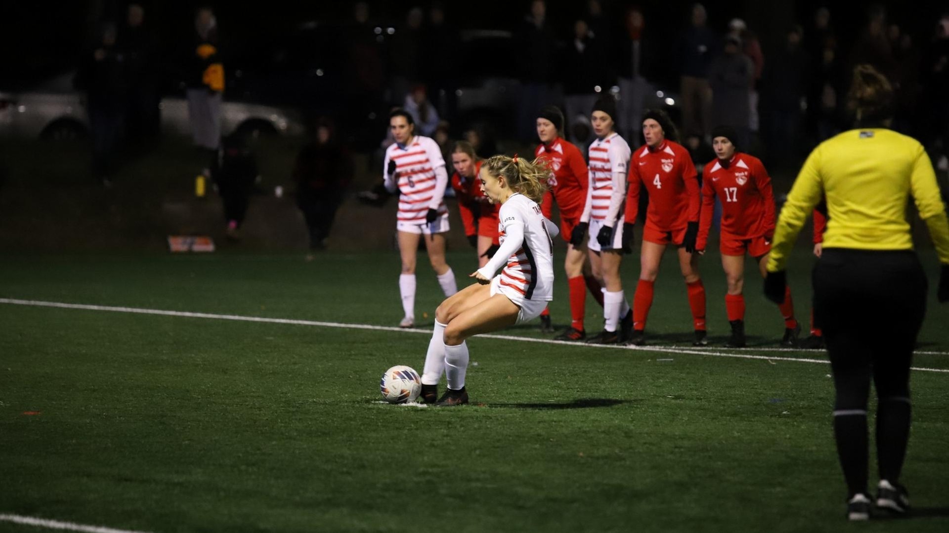 women's soccer player wearing white uniform striking a shot from the penalty mark with players standing behind her
