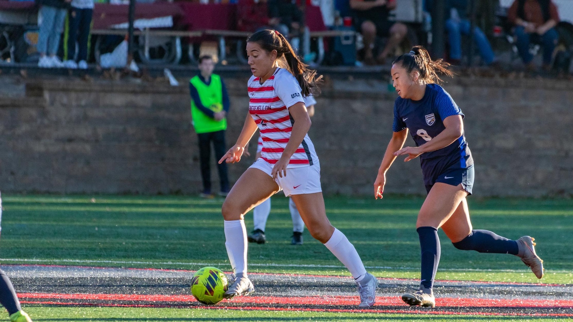 women's soccer player wearing a white and red striped jersey with white shorts dribbles by a defender wearing a blue uniform