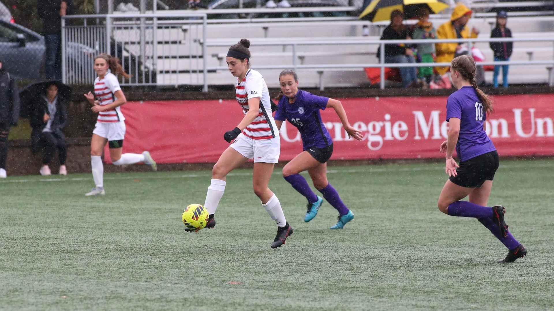 women's soccer player wearing white and red striped jersey and white shorts dribbles the ball with two players in purple uniforms chasing her