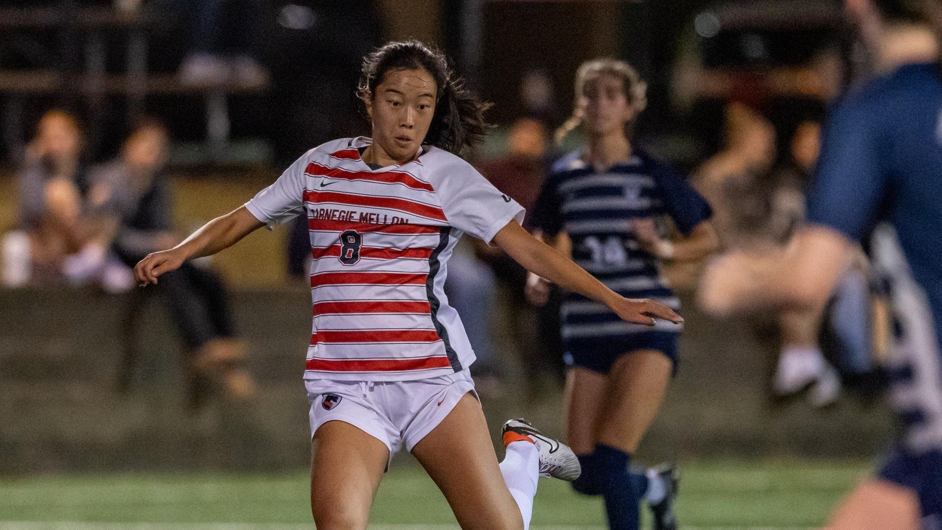 women's soccer player wearing a white and red striped jersey looks down while preparing to kick a ball