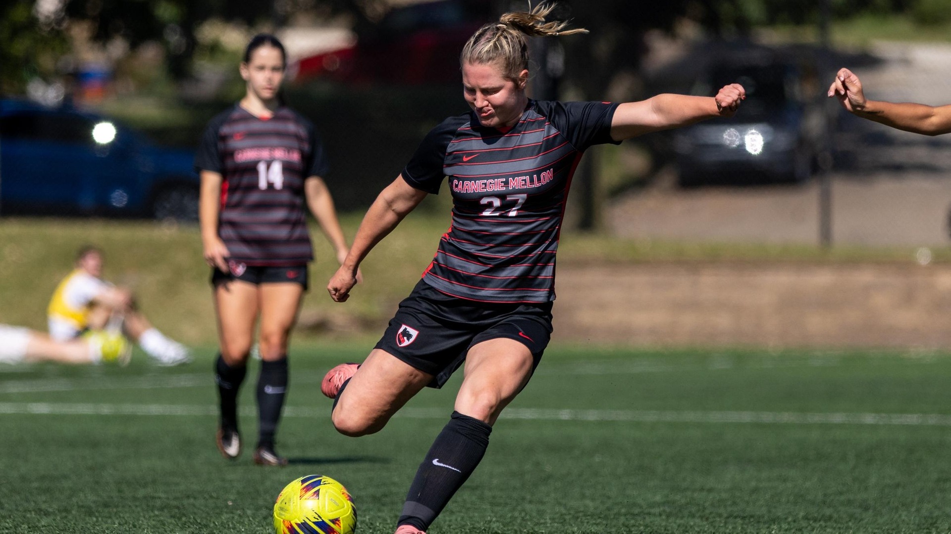 women's soccer player wearing a black jersey and shorts looks down to kick the ball with her right foot