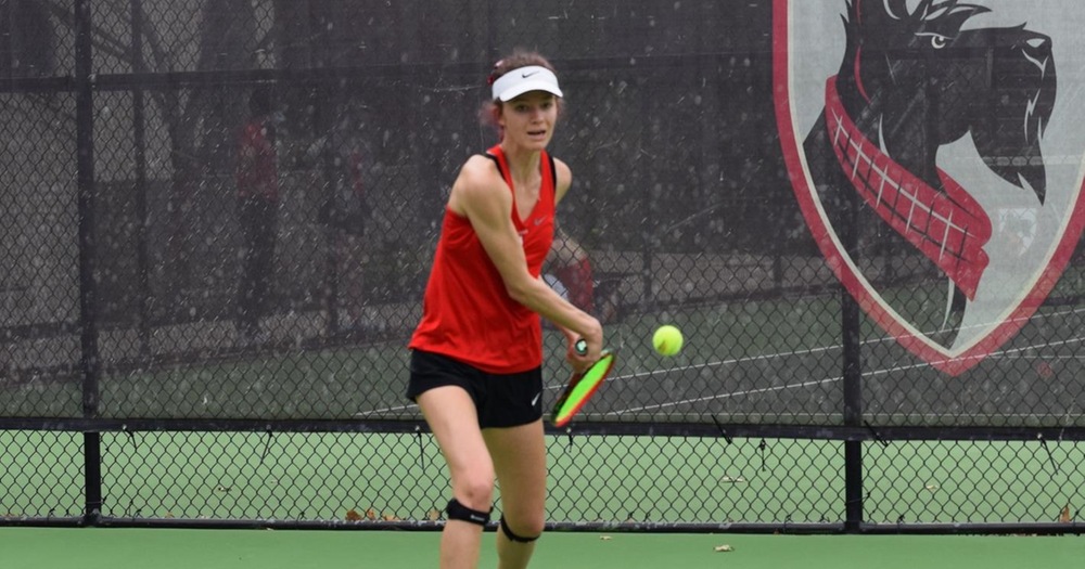 Women's tennis player with red tank, black shorts, and white visor about to hit a backhand shot
