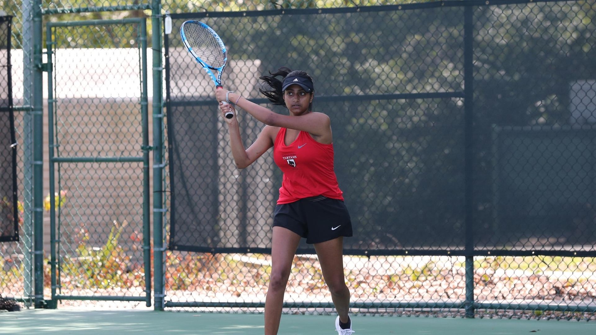 women's tennis player wearing a red top and black skirt with a black visor swings the racquet