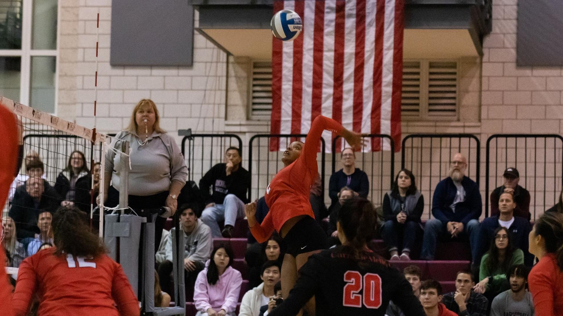 women's volleyball match with a player jumping in the air to hit the ball with her left arm and fans sitting in the background