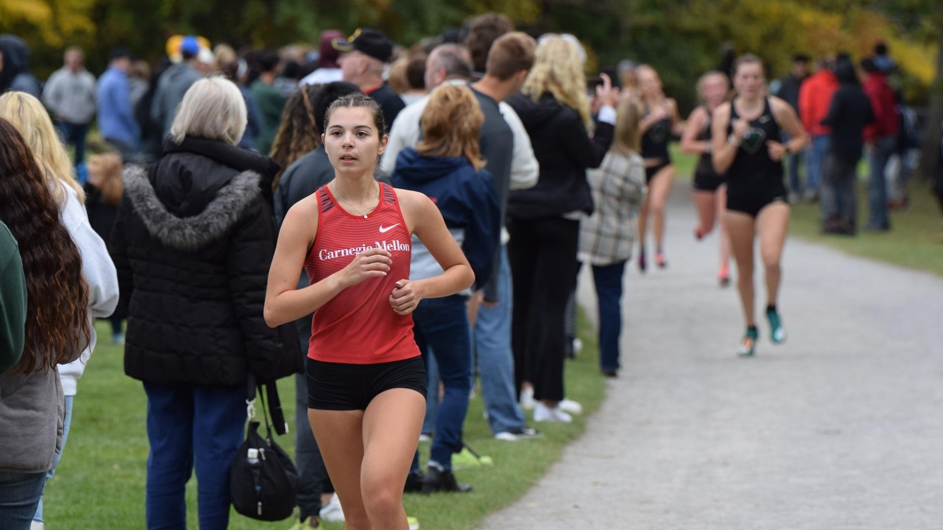 women's cross country runner wearing red jersey and black shorts races on the trail with fans watching behind her