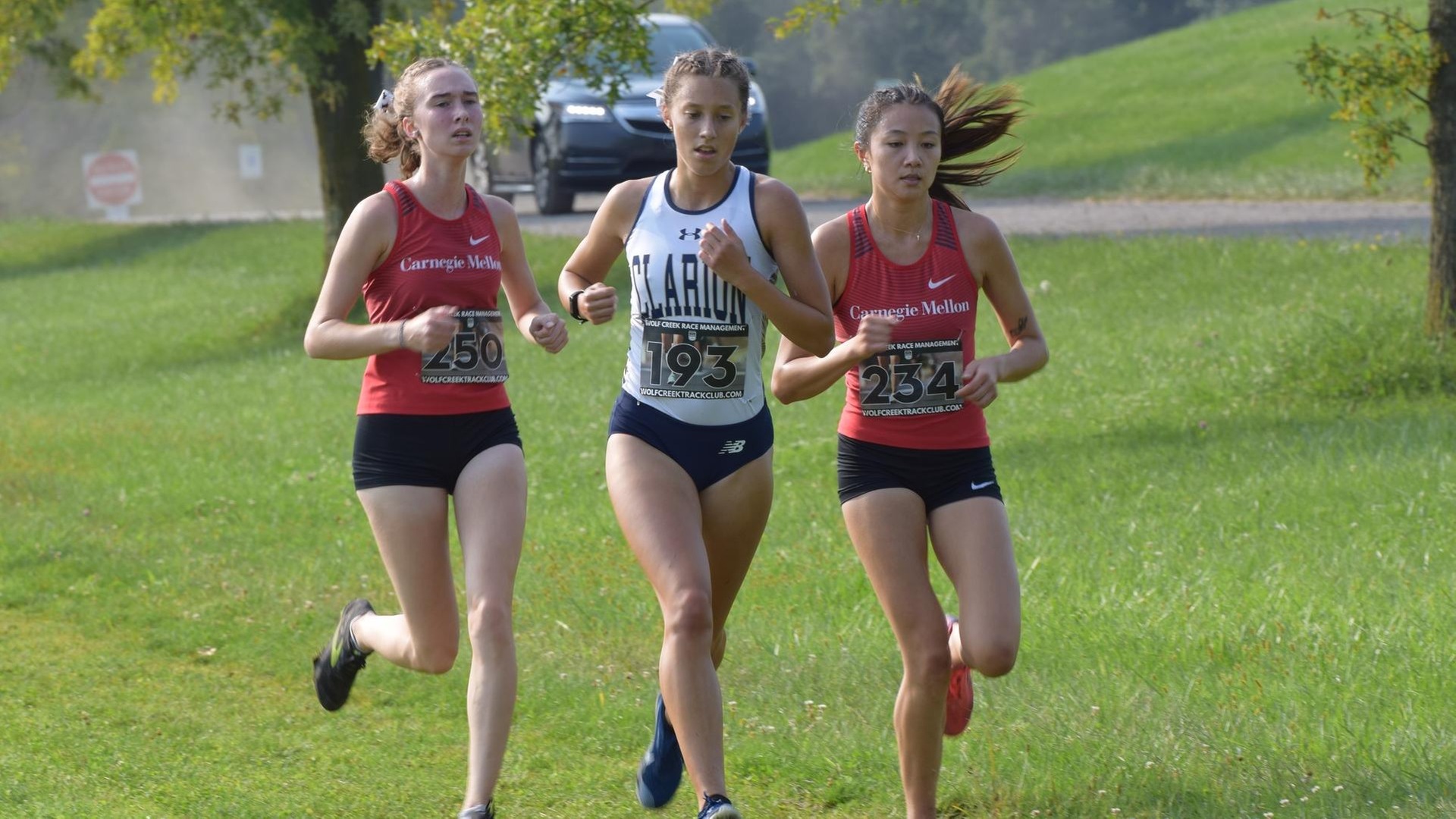 three women's cross country runners on a trail with green grass and a car in the background
