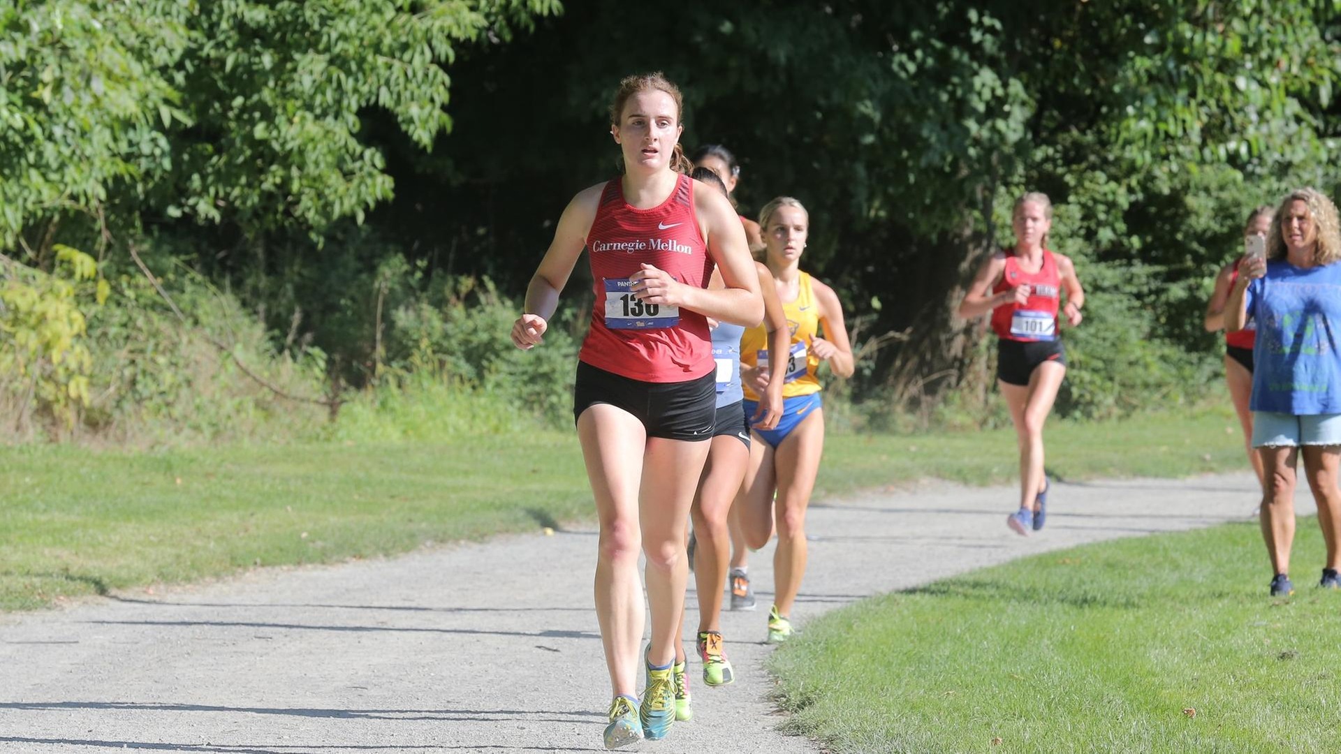 women's cross country runner wearing a red shirt and black shorts leading a group of runners on a trail