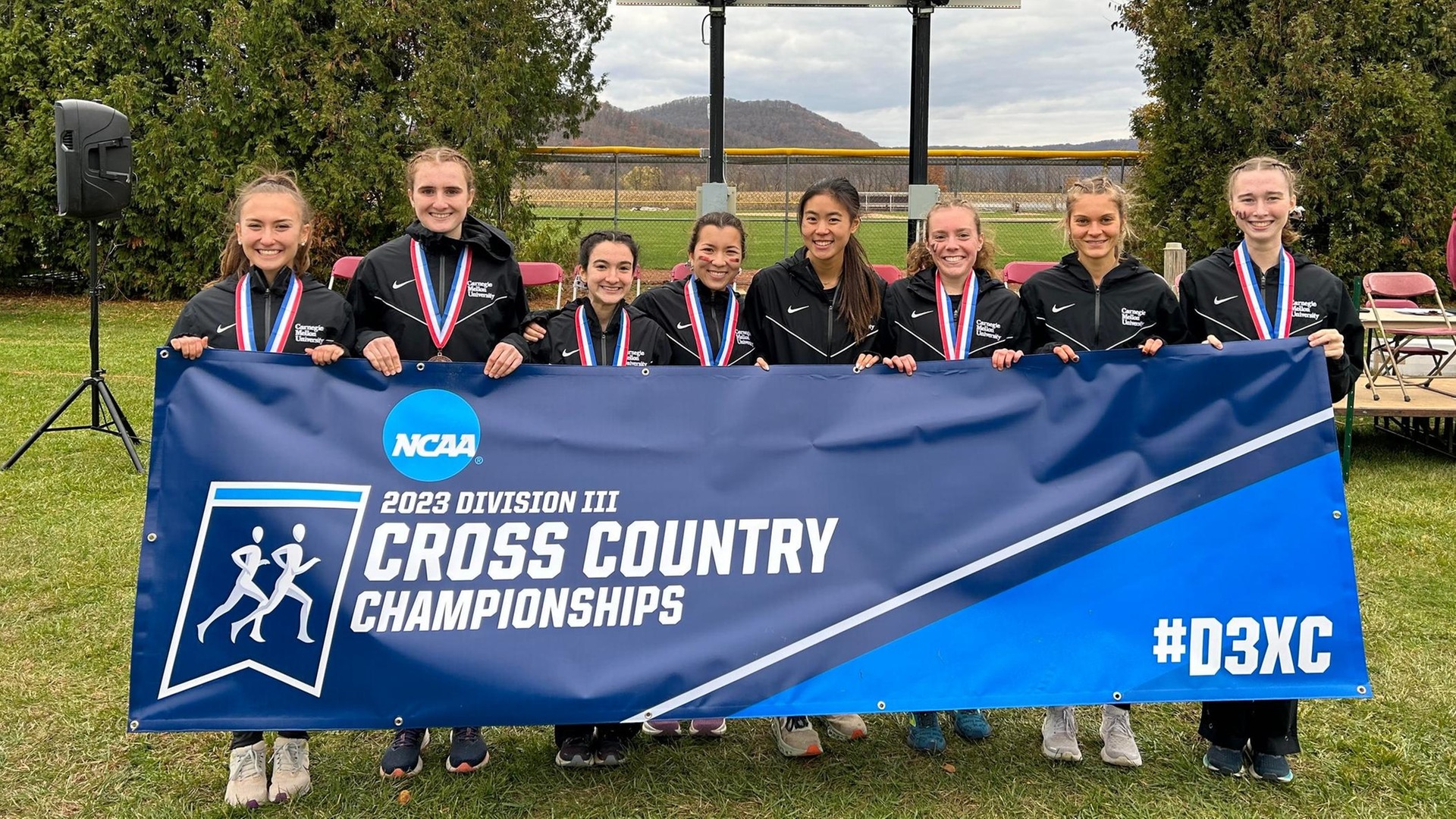 group of women wearing black track suits holding a banner that is blue and says 2023 Division III Cross Country Championships